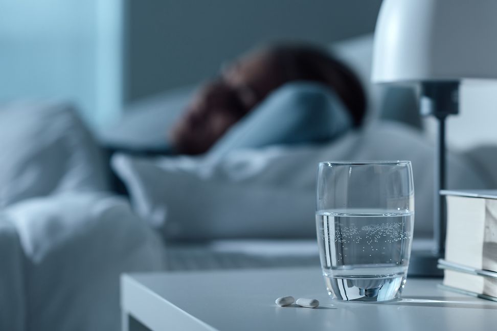 Glass of water and a pill next to a bed with a sleeping person inside