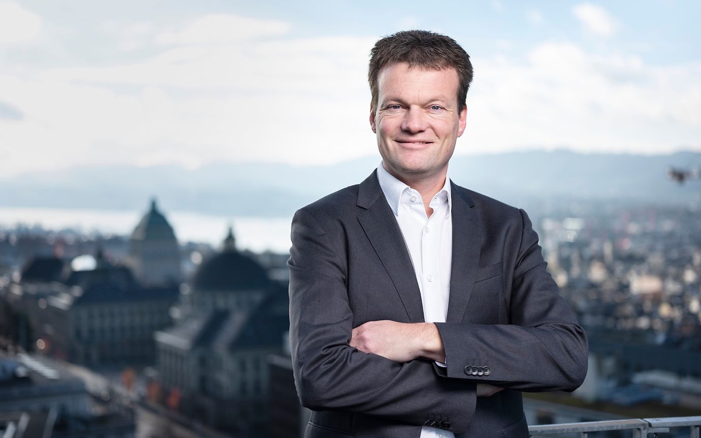 Reto Knutti, auf einer Terrasse stehend mit dem Zürichsee und der Universität im Hintergrund, blickt in die Kamera.