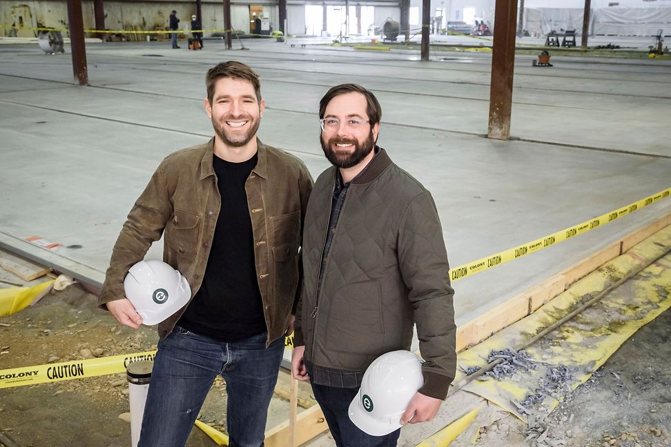 Two young men with hard hats in hand in a warehouse
