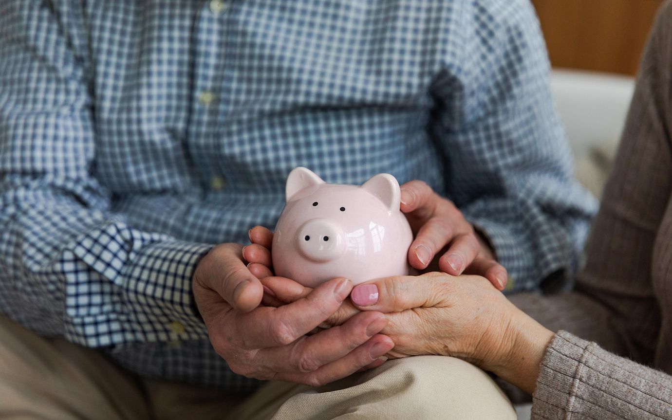 A pink piggy bank, being held by male hands intertwined with female hands that are painted with pink nail varnish.