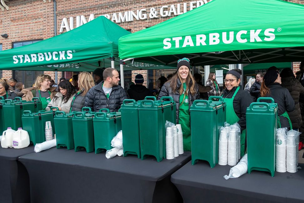 A group of young people pour coffee from large warming containers under Starbucks umbrellas.