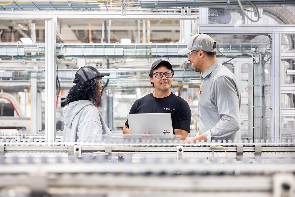 Three people are talking around a laptop in an industrial production hall. 