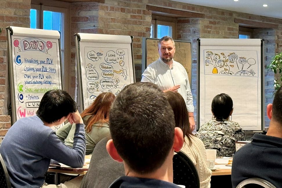 Man with a beard presents with three flipcharts to a group of young people.