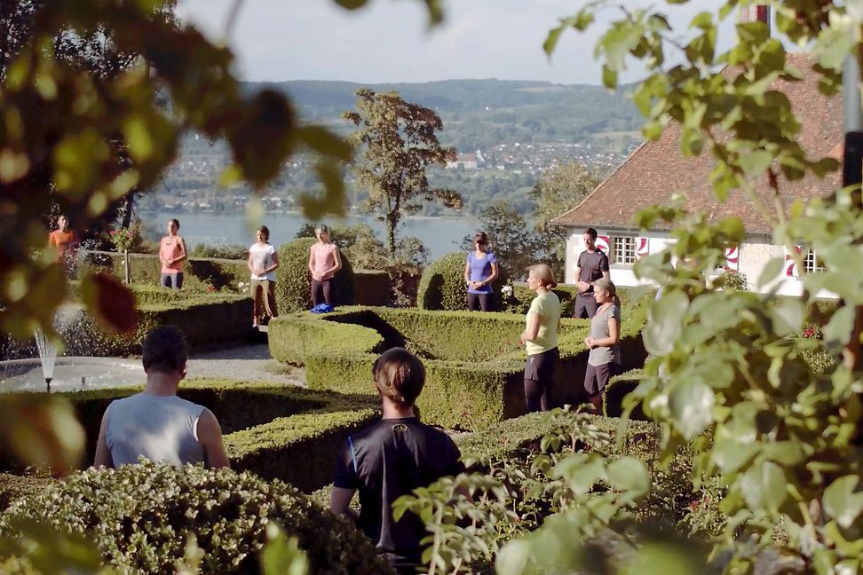 A diverse group of people stand in the rose garden of a castle and do meditation exercises in the sunshine.