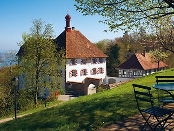 Kleines Schloss mit rot-weissen Fensterläden liegt eingebettet in Bäume und Wiesen oberhalb eines Sees im Frühling. 