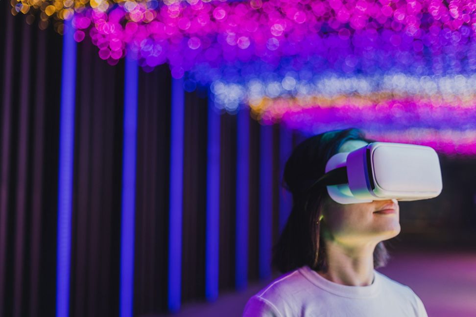 A person wears virtual reality glasses against a backdrop of pink and purple lighting.