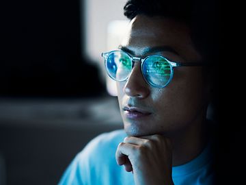 Nahaufnahme eines Mannes, Kinn auf Hand, dessen Computerbildschirm sich in seiner Brille spiegelt.
