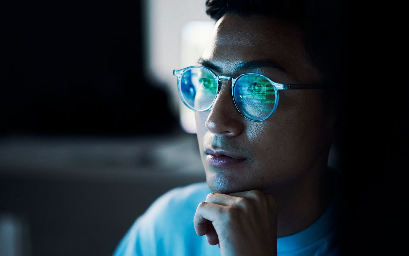 Nahaufnahme eines Mannes, Kinn auf Hand, dessen Computerbildschirm sich in seiner Brille spiegelt.