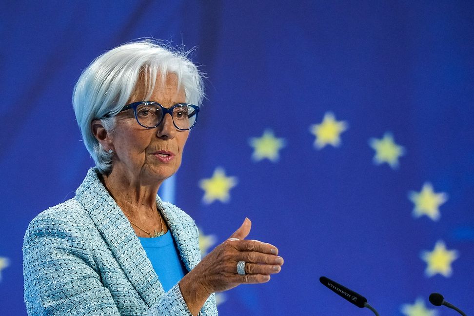 A woman in a suit and glasses gesticulates at the microphone, the EU flag in the background.