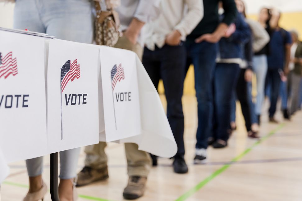 People are waiting in a gymnasium in front of a table marked "Vote" and an American flag.