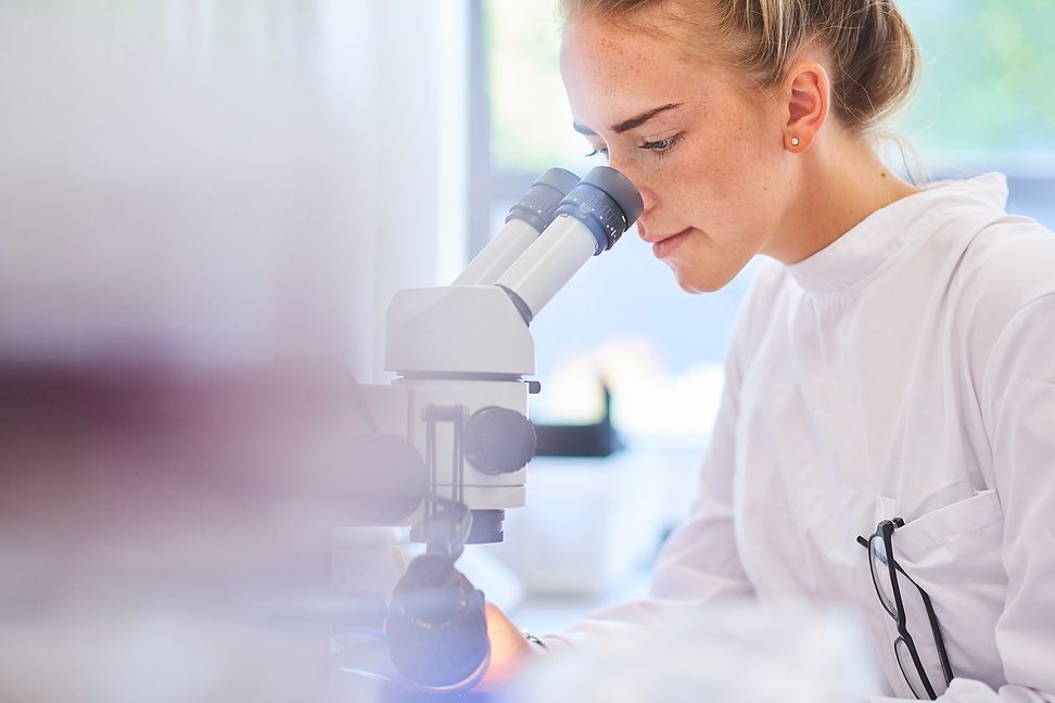 A young woman in a white lab coat looks through a microscope in a laboratory environment.