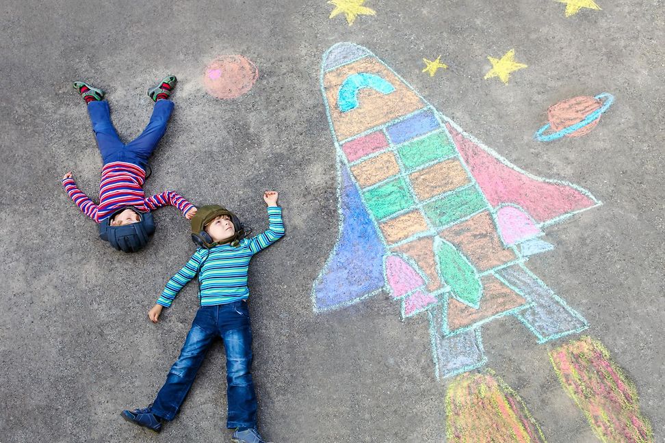 Two children lie on their backs on a concrete floor, holding a brightly coloured rocket they have painted on the floor