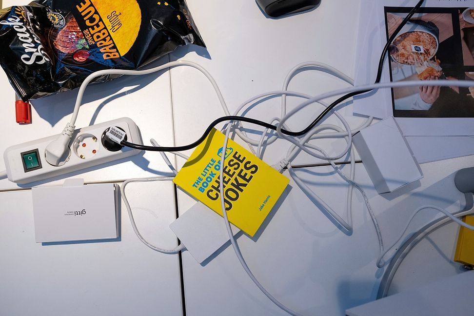 Auf einem Tisch, aufgenommen von oben, liegt ein Witzbuch mit gelbem Buchdeckel neben einer Steckerleiste