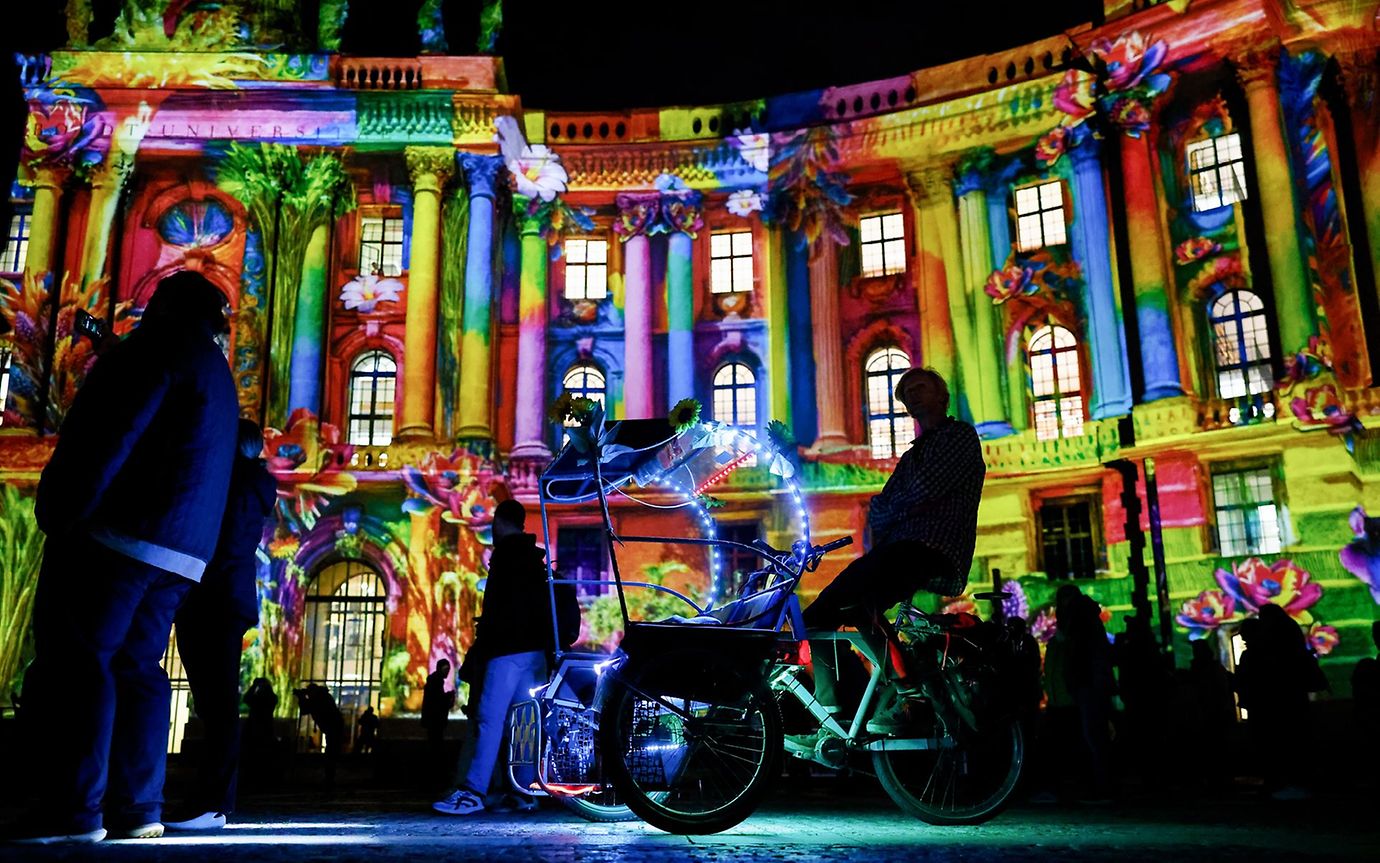 Colourful lights on a large official building in classical style. In the foreground you can see dark silhouettes of many people.