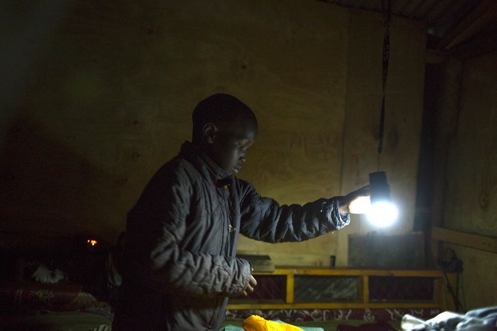 A man stands in a simple, dark shelter holding a light bulb.