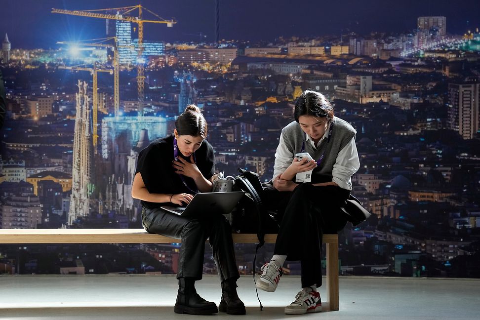 A man and a woman are sitting on a bench looking at their laptops and mobile phones. In the background the city skyline by night