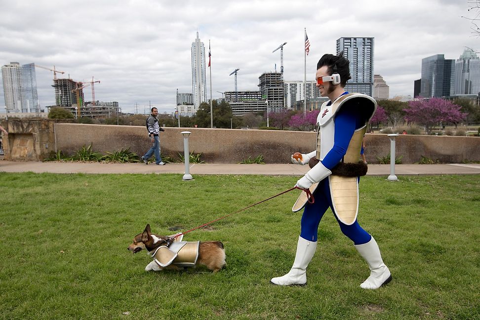 A man in a futuristic costume walks his dog in a matching costume. In the background is the skyline of a large city.