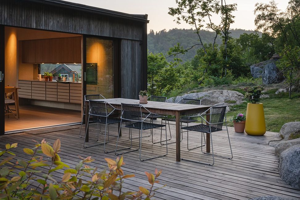 Modernes Holz-Glashaus lädt mit warmem Licht und offener Terrassen-Tür. Vorne auf Holzterasse steht grosse Esstisch.