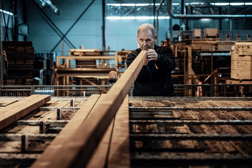 Mann in einer Produktionshalle hält einen langen Balken in der Hand uns blickt prüfend die Holzkante entlang .