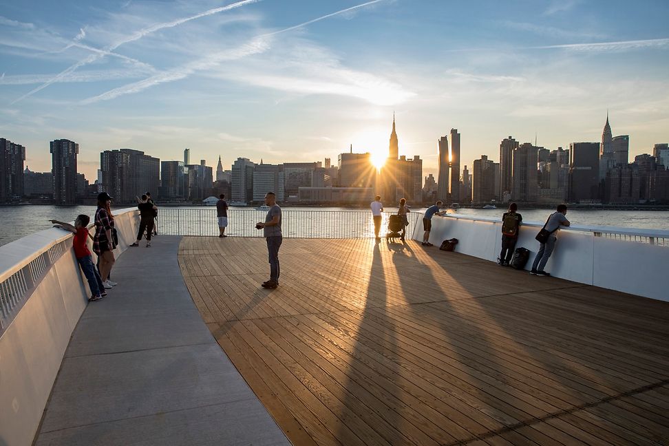 Gegen die Abendsonne stehen fünf Individuen auf einem Holzsteg am Ufer eines Gewässers, Hintergrund urbane Skyline