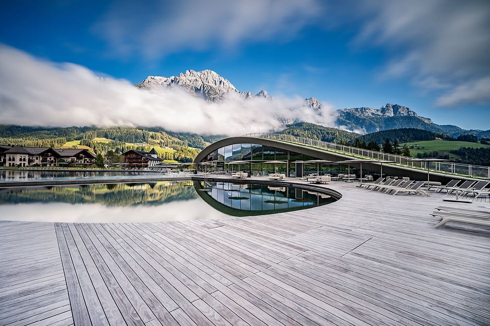 Vor Alpenpanorama und gegen einen blauen Himmel erstreckt sich ein grosszügiger Holzsteg und eine moderne Fussgängerbrücke. 