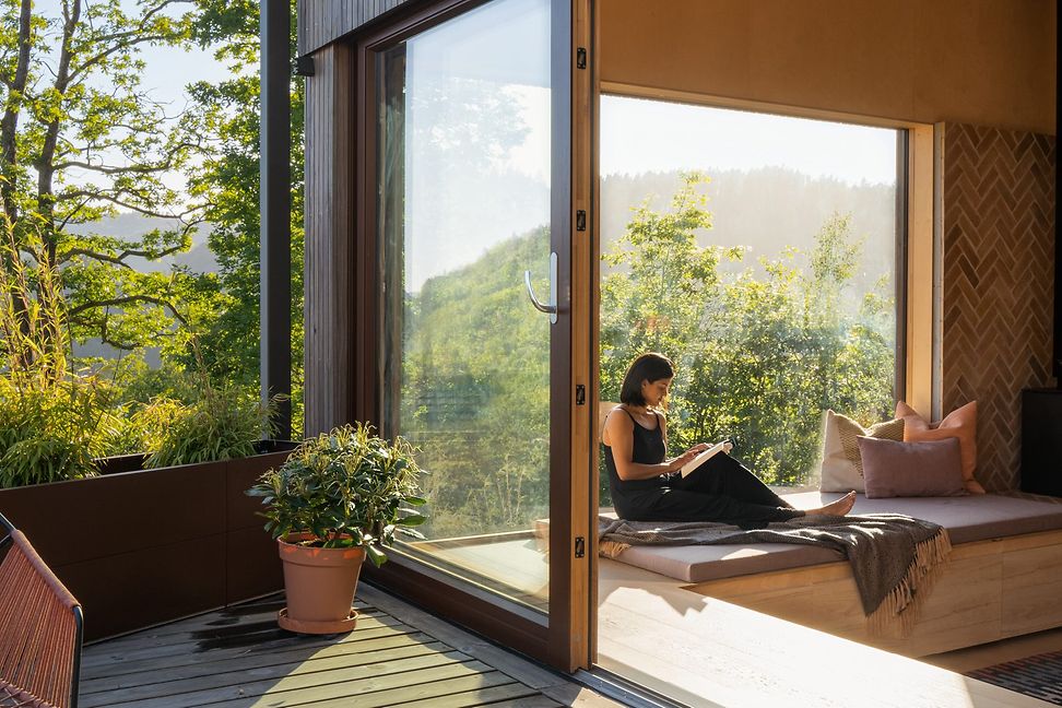 A light-filled room that appears to be part of a modern glass and wood house. A woman is reading in a window seat.