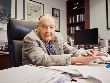 An elderly businessman sits at a desk surrounded by books, papers, pictures and a computer screen.