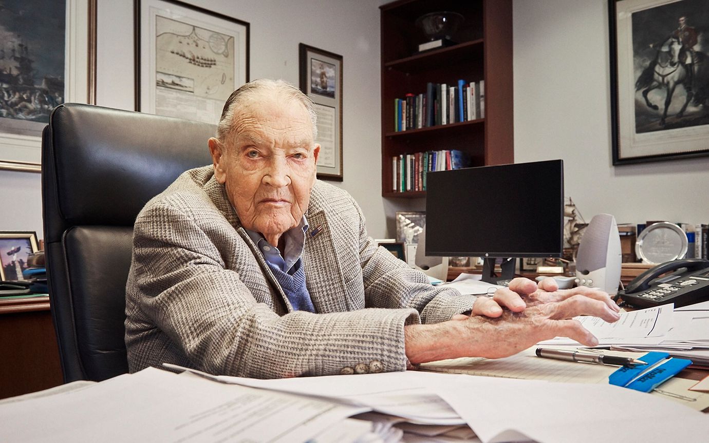 An elderly businessman sits at a desk surrounded by books, papers, pictures and a computer screen.