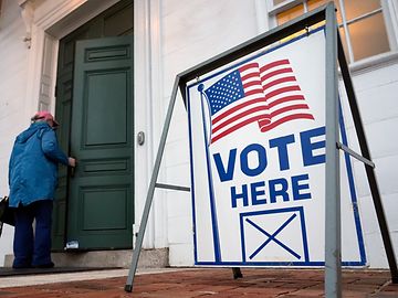 Eine Person betritt ein Haus mit einer grünen Tür, vor der ein Schild mit der Aufschrift "Vote here" und einer US-Flagge steht.