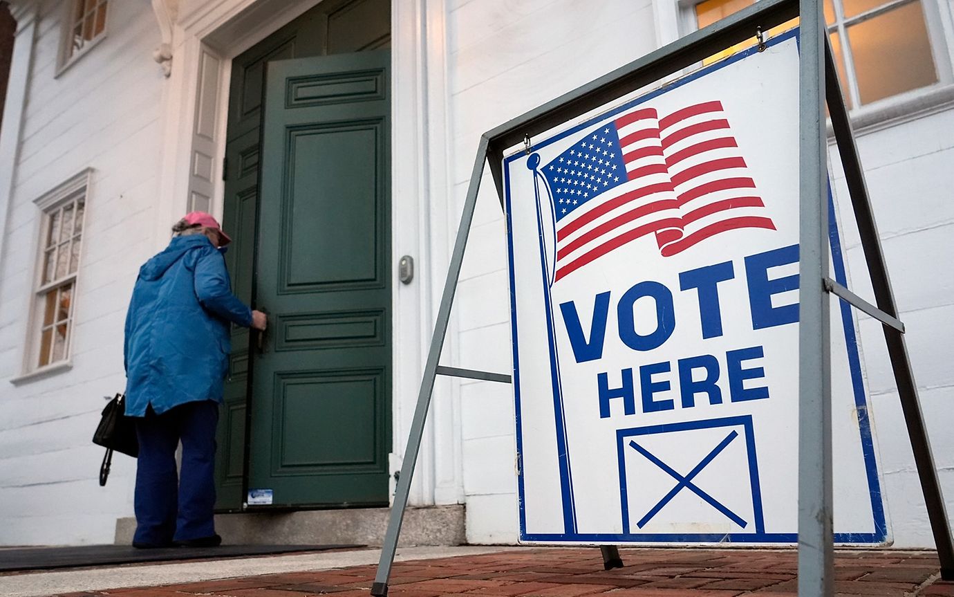 Eine Person betritt ein Haus mit einer grünen Tür, vor der ein Schild mit der Aufschrift "Vote here" und einer US-Flagge steht.