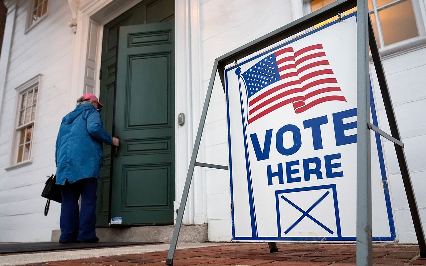 A person enters a house with a green door in front of which is a sign saying 'Vote here' and a US flag.