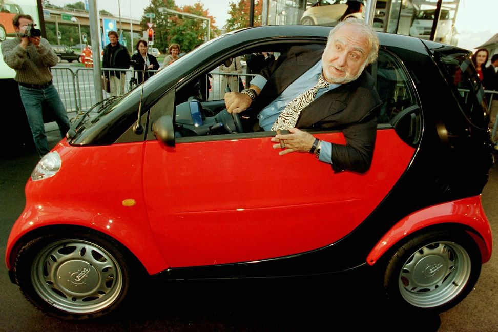 Ein älterer Herr lehnt sich aus dem Fenster eines roten sehr kleinen Autos, während die Leute zuschauen und fotografieren.