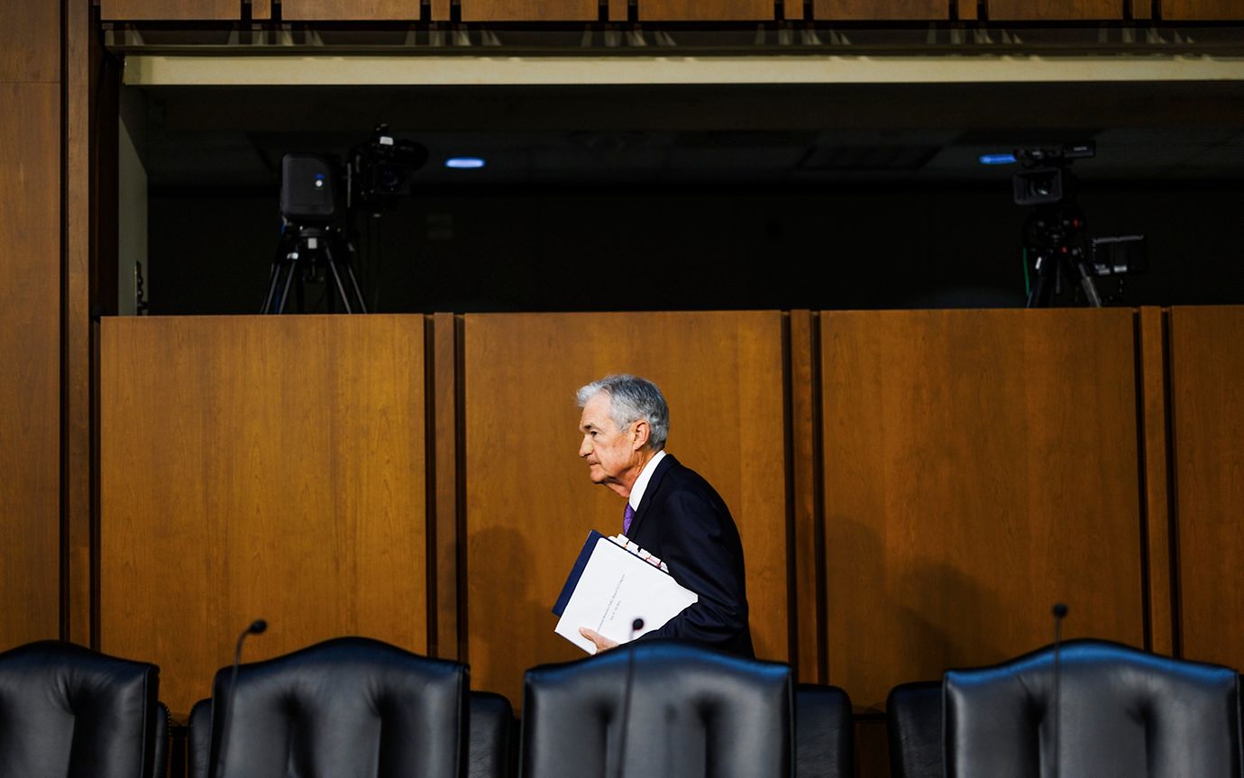 Elderly man in a suit with folders in his hands enters a room with chairs with microphones and a video production room behind 