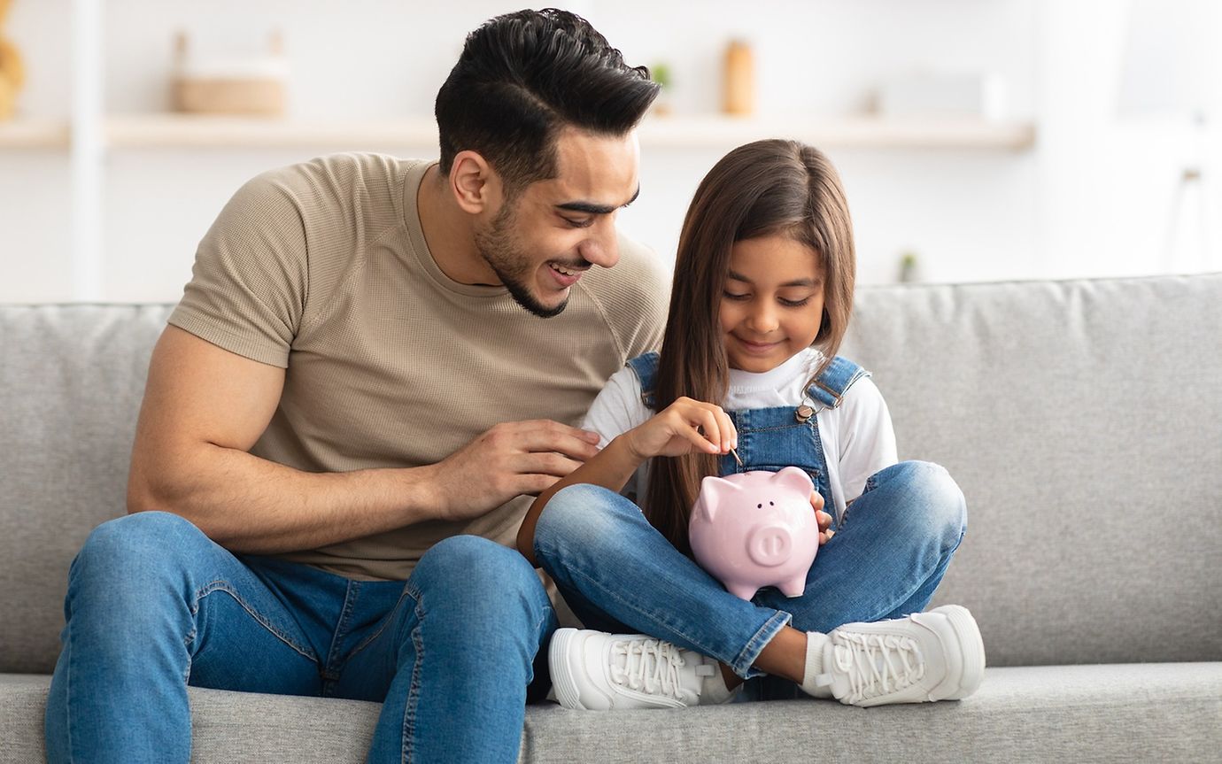 A man and a child are sitting on a sofa in a domestic setting, putting something into a pink clay pig.