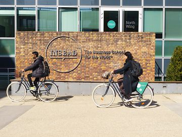 Two people on bicycles pass a brick wall in front of a building with the word “INSEAD” on it.