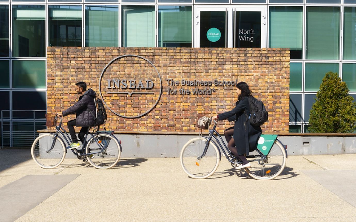 Two people on bicycles pass a brick wall in front of a building with the word “INSEAD” on it.