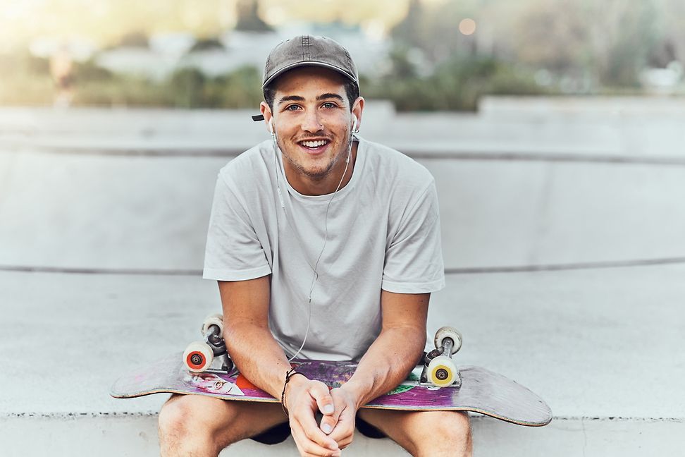 Ein junger Mann sitzt mit einem Skateboard auf dem Schoss und lächelt in die Kamera.