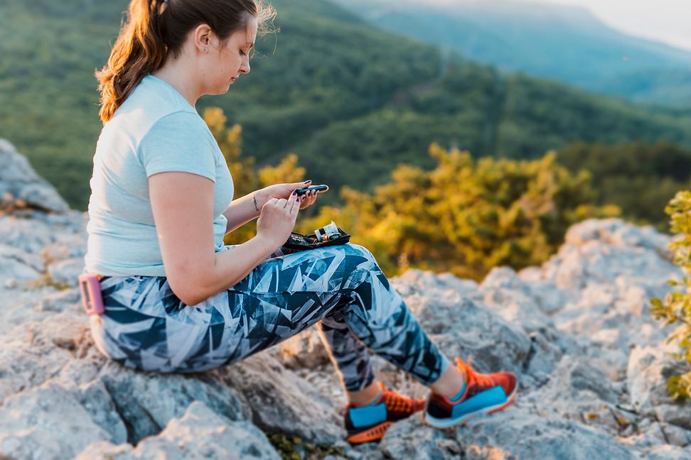 A young woman in sportswear sits on a large rock in a mountainous landscape using an insulin pen.