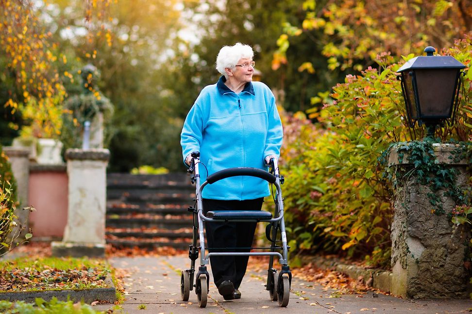 A white-haired elderly woman walks with a walking frame in an autumnal park.