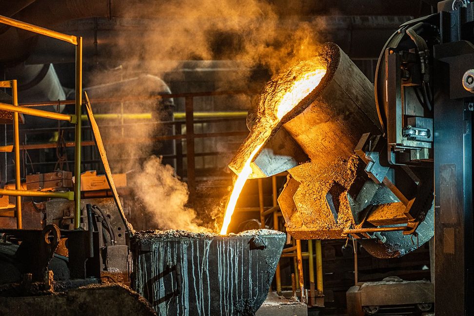 In an industrial factory, molten metal is poured into a massive vessel, causing smoke to rise.