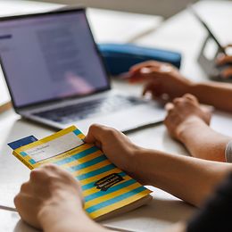 Ein Laptopbildschirm und ein Schulheft in den Händen von Personen, von der Seite gesehen.