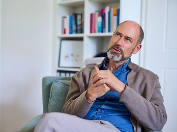 A middle-aged man with a bald head and a grey beard is sitting in an armchair