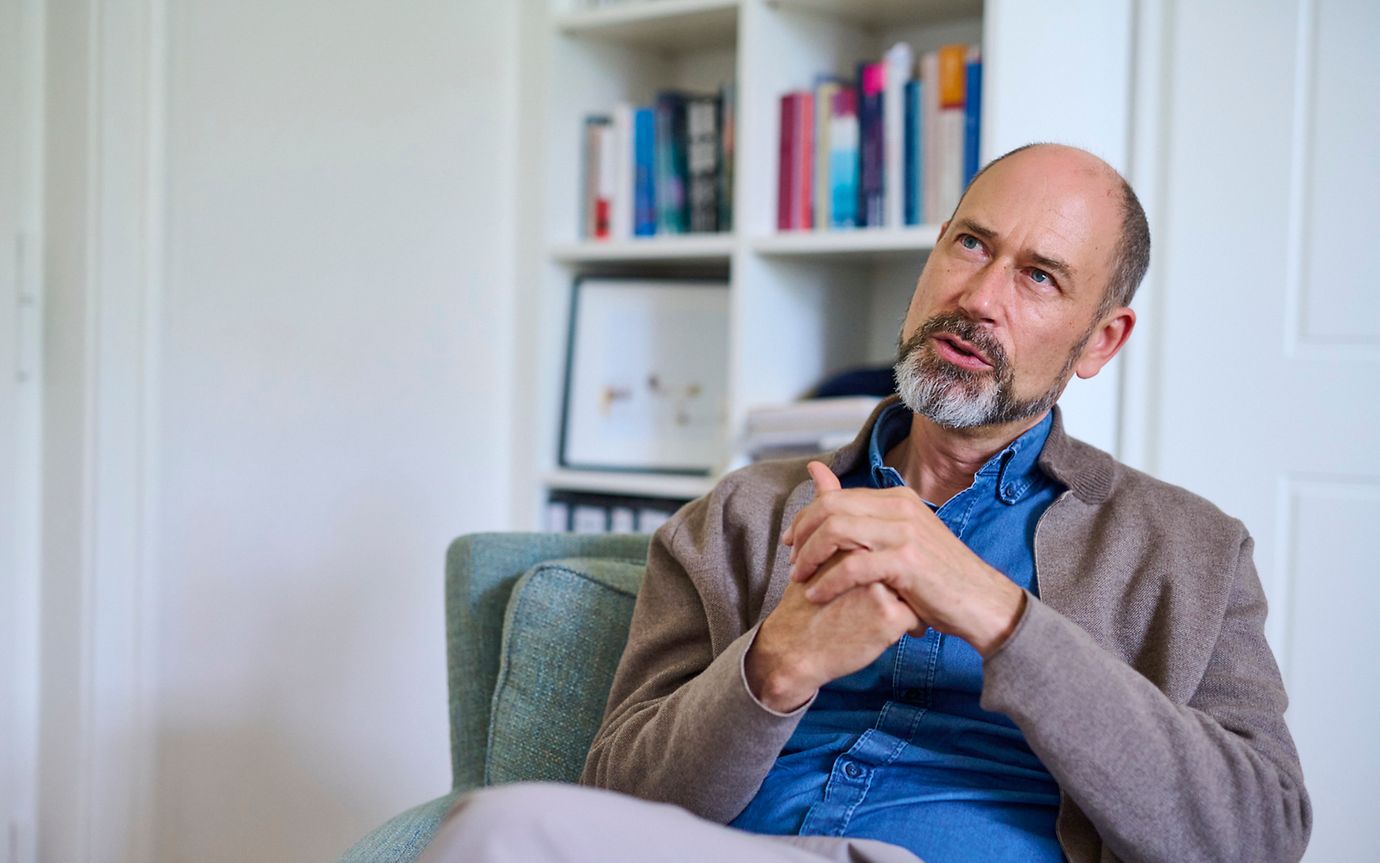 A middle-aged man with a bald head and a grey beard is sitting in an armchair