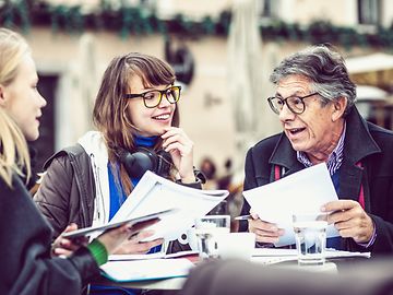 An older man is talking to two younger women, all holding documents, in an outdoor café.