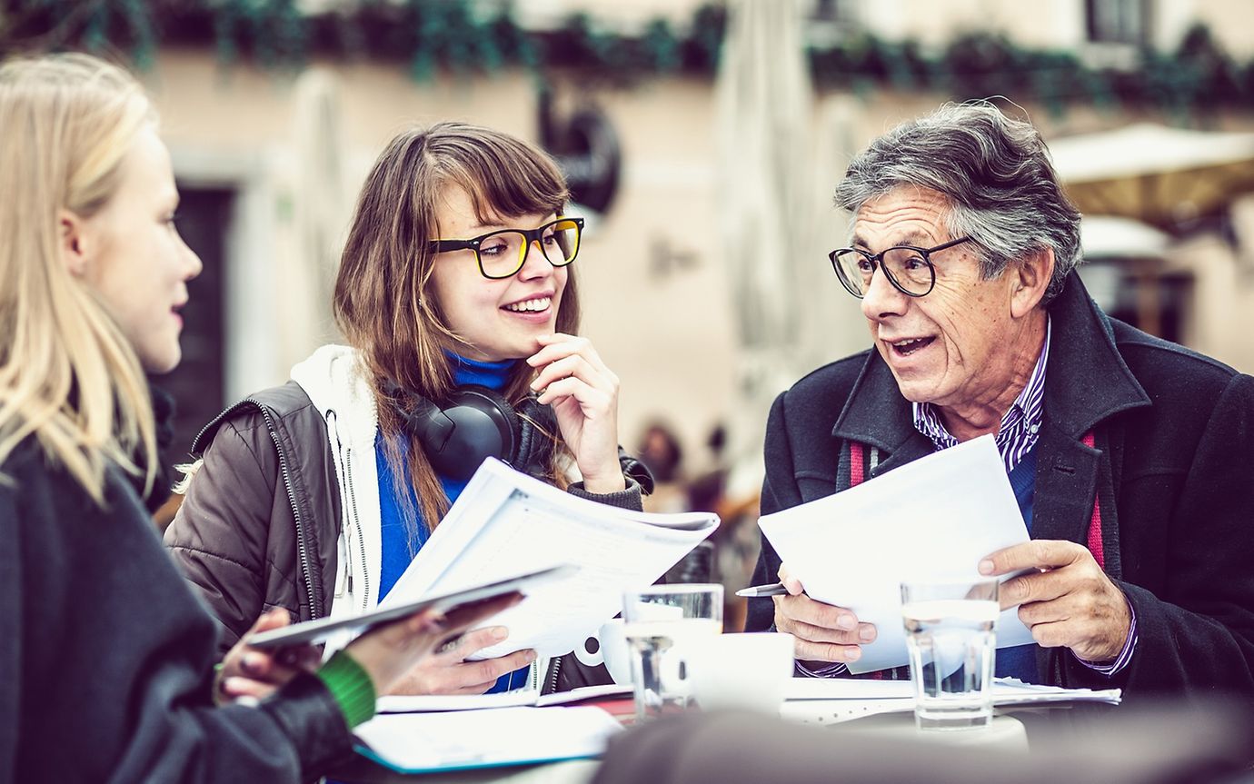 An older man is talking to two younger women, all holding documents, in an outdoor café.