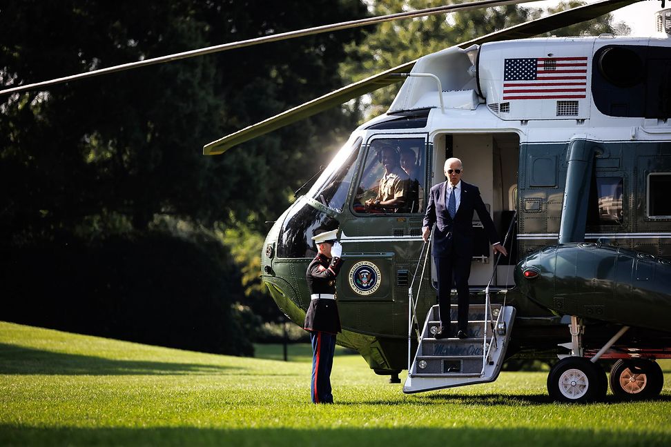 An elderly man in a suit steps out of a helicopter with a US flag on a green field as a soldier greets him