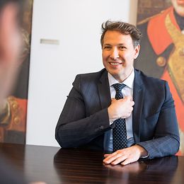 A man in a suit and tie in an office environment talks and touches his tie