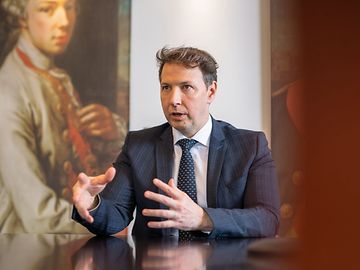 A man in a suit and tie in an office environment talks and gestures with his hands