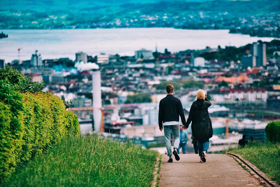 A young couple holding hands walk along a pavement surrounded by greenery towards a city on a lake in the background.