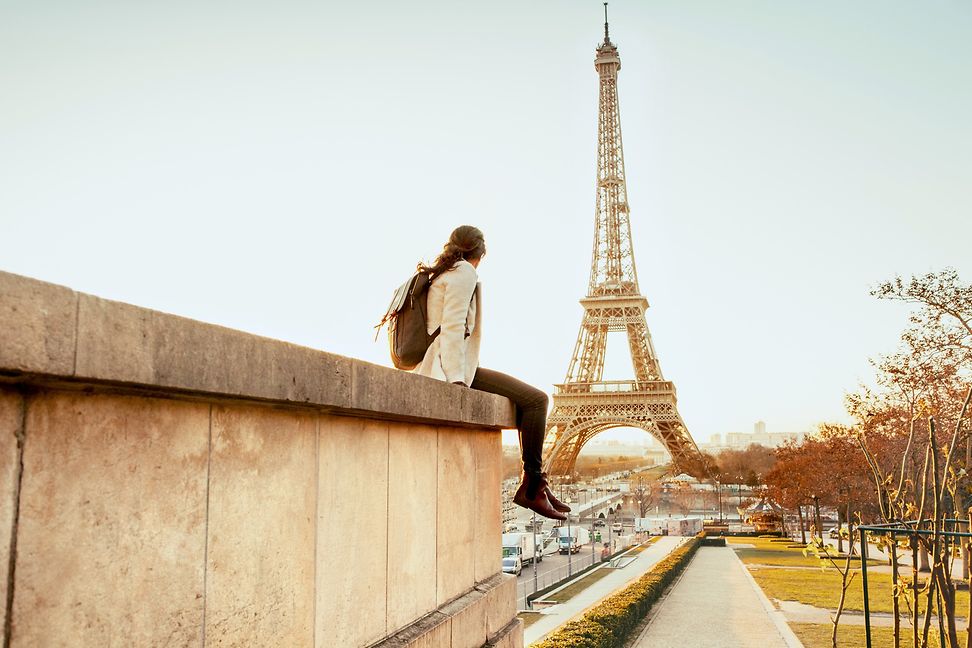 A woman wearing a black rucksack, white jacket and brown trousers is sitting on a stone wall,